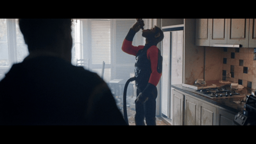 Medium long shot of Williams in the aforementioned "space-age-fat-sucking-vacuum-suit" chugging a beer in a messy kitchen as his best friend looks on from the foreground in silhouette