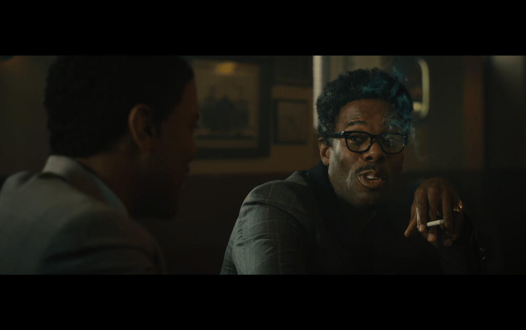Close-up of Bayard Rustin sitting at a bar talking to Elias Taylor (who has his back to the camera in the foreground) while he smokes a cigarette