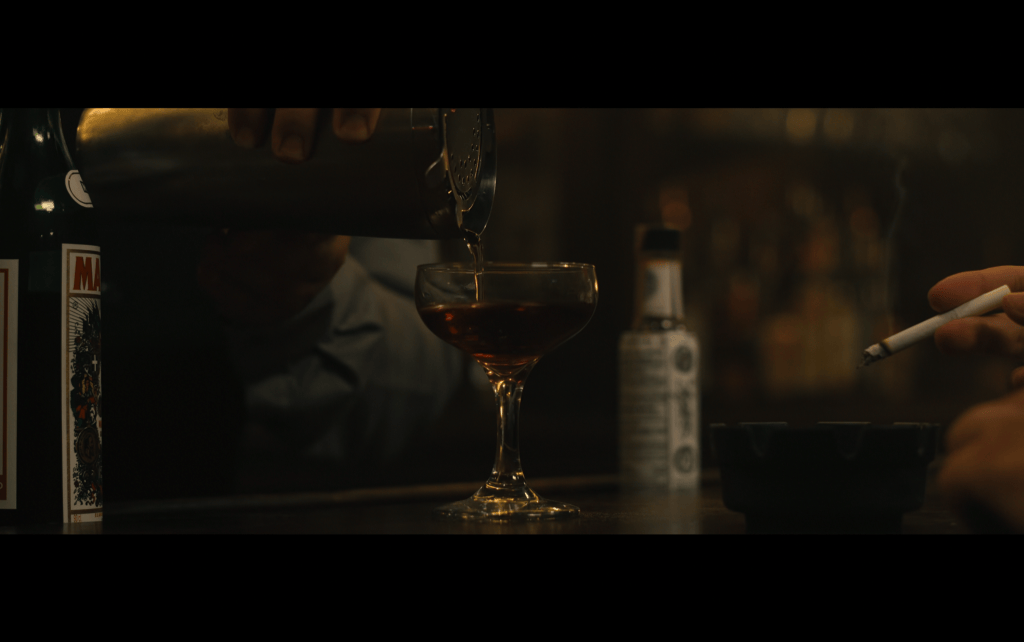 Close-up of a coupe glass being filled from a shaker in front of a bottle of Angostura bitters