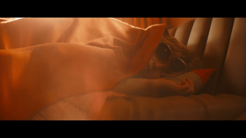 Medium close-up of a clearly hungover Kara sleeping under a red blanket while still wearing a pair of askew sunglasses