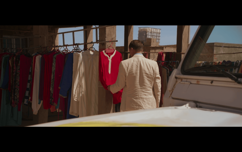 Exterior daytime medium shot of David examining a red shirt in the local style that hangs on a rack with his back to the camera