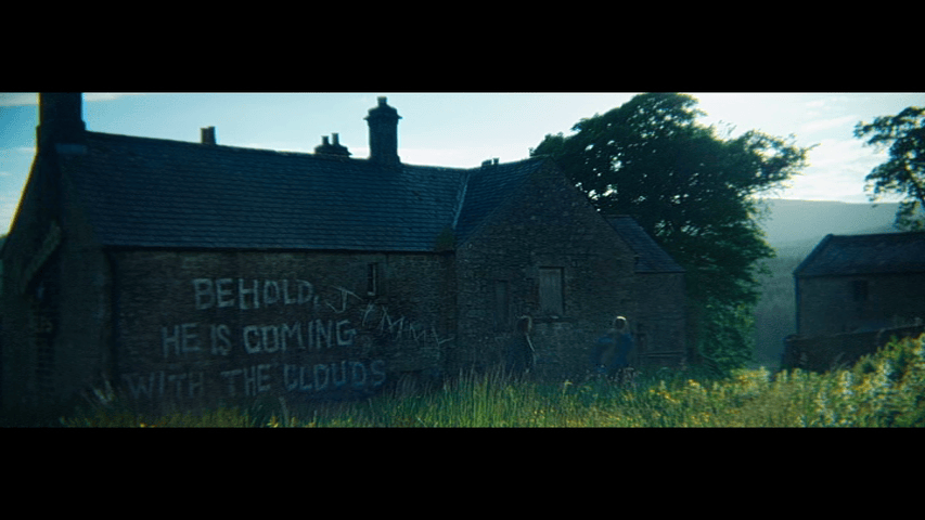 Long shot of Spike and his mother walking in front of a stone building that has the words "Behold He Is Coming With the Clouds" written on its wall in large white letters and the name "Jimmy" appended to the message in different handwriting
