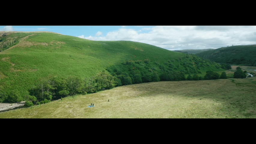 Extreme long shot of three figures dwarfed by the cloud shadow moving across the rolling hills around them