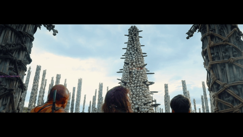 Low-angle shot of Dr. Kelson, Spike, and Spike's mother Isla (Jodie Comer) looking up at a tower of skulls flanked on all sides by pillars of bone