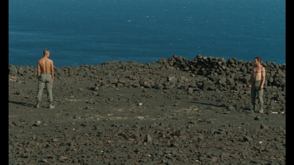 Long shot of Denis Lavant's Galoup and Grégoire Colin's Sentain staring each other down from screen left and right respectively on a rocky terrain in front of a glistening blue sea which occupies the top third of the frame