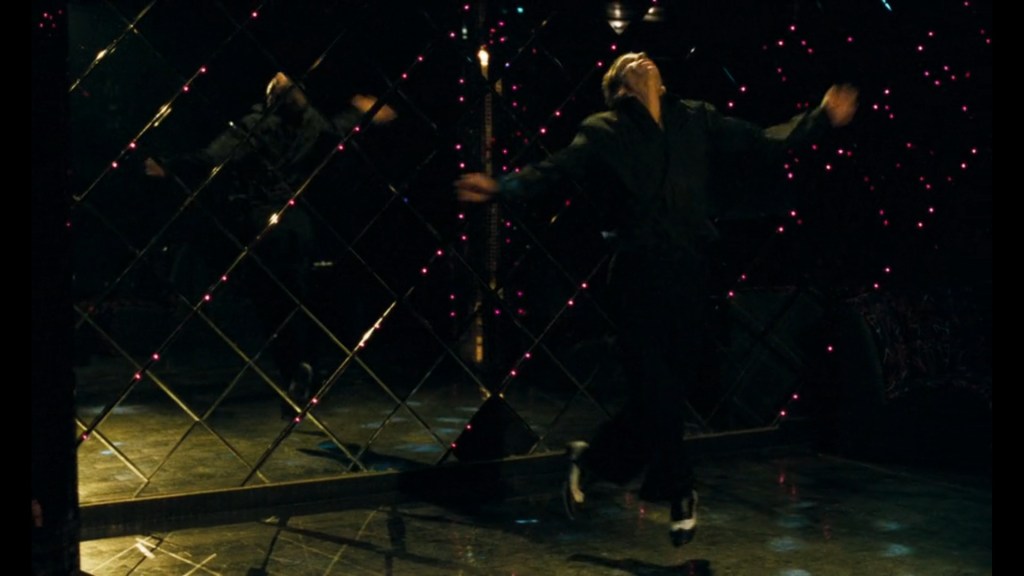 Medium shot of Denis Lavant's Galoup ecstatically throwing himself in the air, arms spread wide, at a discotheque