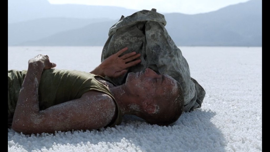 Medium long shot of a parched-looking Sentain lying on a salt flat with only his pack to provide shade