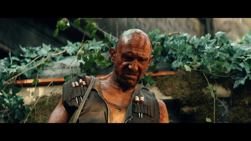 Close-up of Dr. Kelson in front of a vine-covered train car, wearing a vest festooned with poison darts