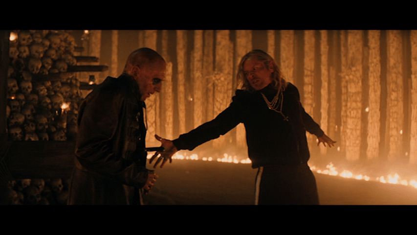 Medium close-up of Sir Lord Jimmy Crystal stabbing Dr. Kelson as he stands in front of his tower of skulls, a line of fire behind them