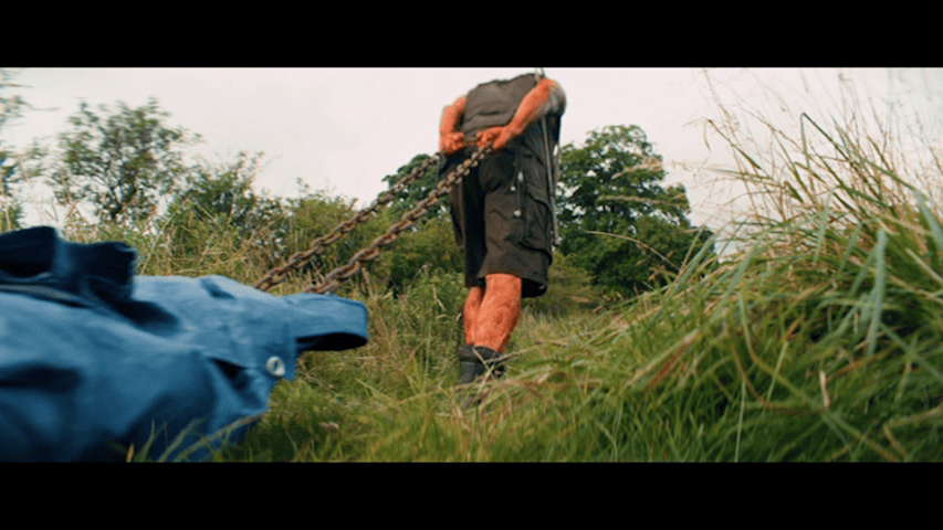 Medium long shot of Dr. Kelson from behind walking across the frame from left to right, dragging a body covered in a blue tarp behind him
